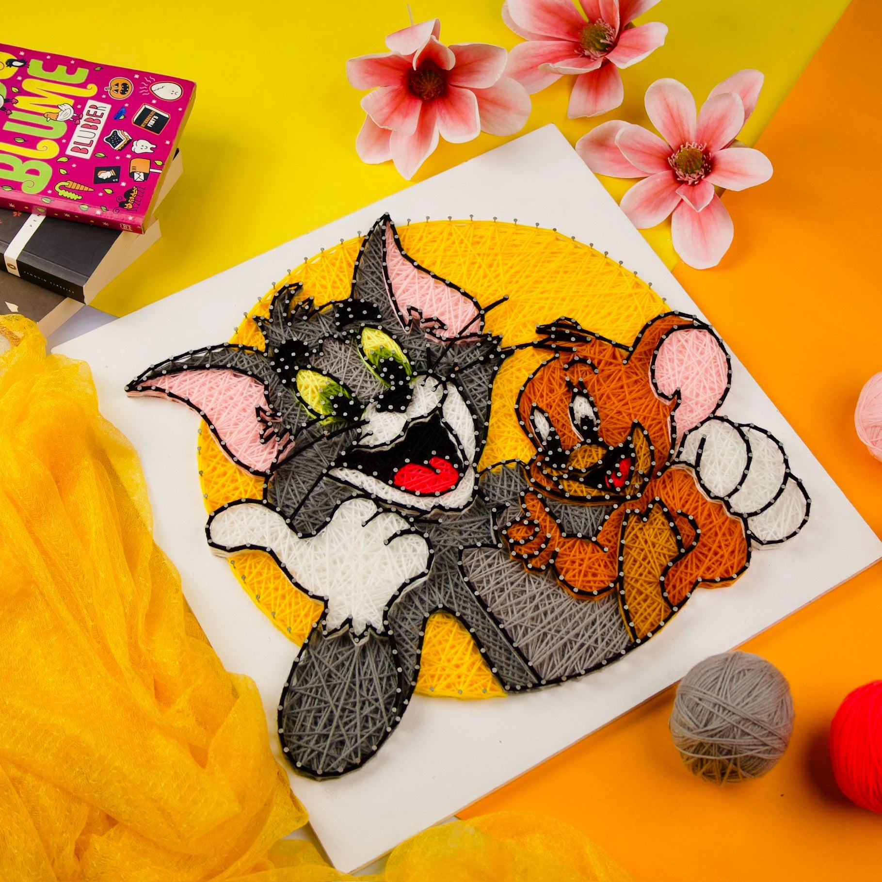 A colorful string art depiction of Tom and Jerry against a yellow background, surrounded by yarn balls and decorative flowers.